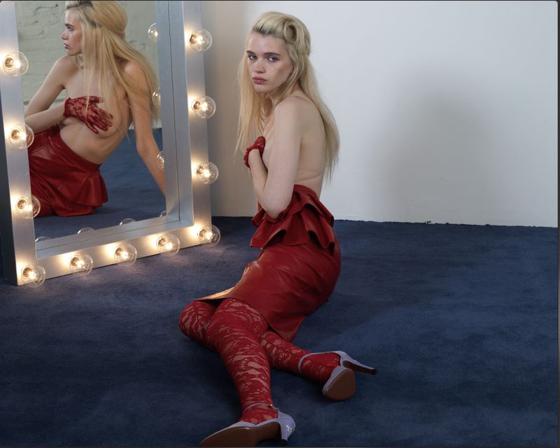 Blonde woman with red gloves and thigh-high boots poses next to a lightbulb mirror in a photo studio against a blue carpet and white wall.