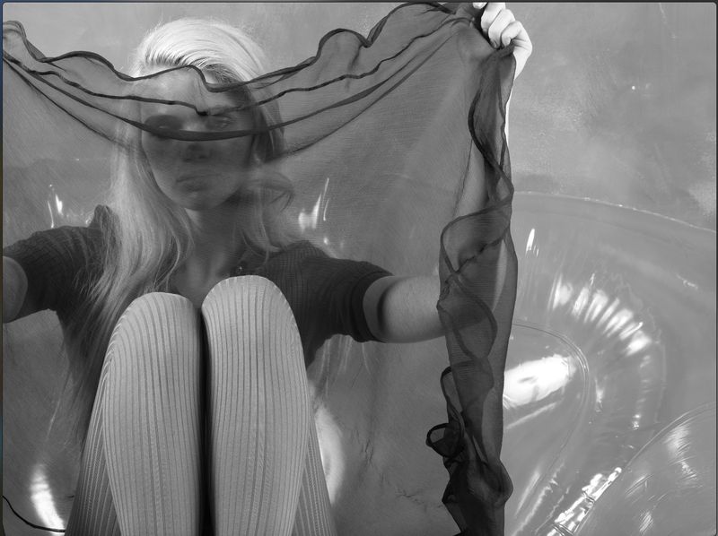 Monochrome studio shot of a woman with long blonde hair sitting with knees up and holding a sheer black fabric.