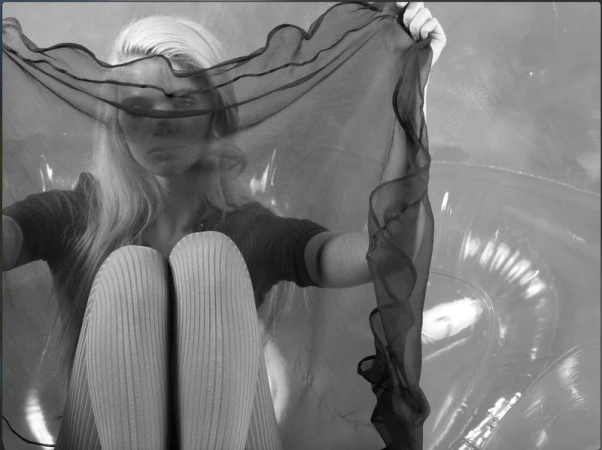 Monochrome studio shot of a woman with long blonde hair sitting with knees up and holding a sheer black fabric.