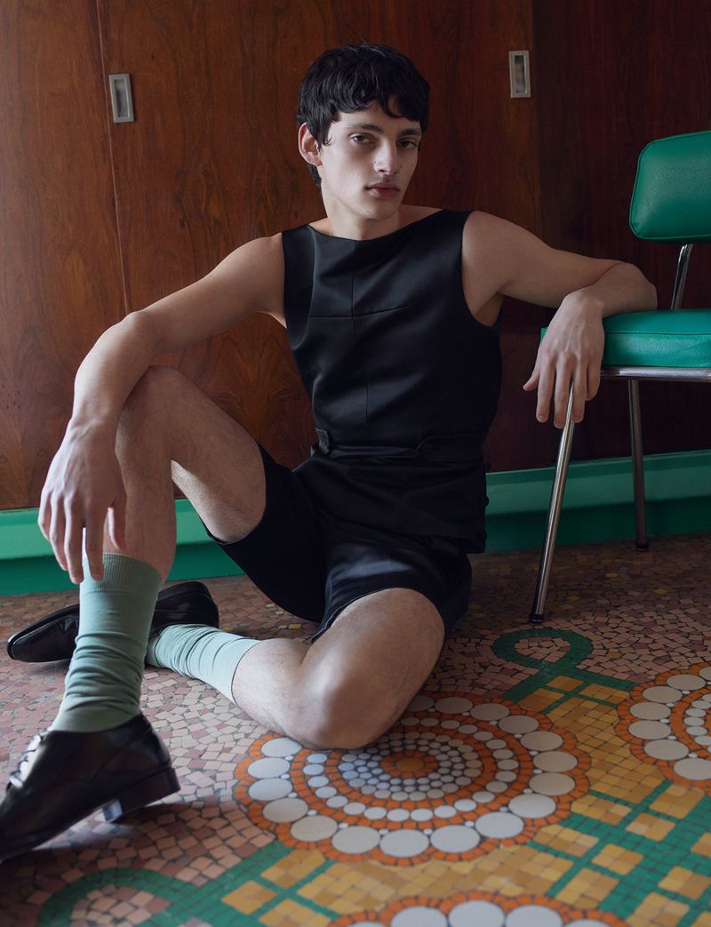 Model wearing a black sleeveless top, shorts, sage green socks, and black leather shoes seated on a patterned mosaic floor.