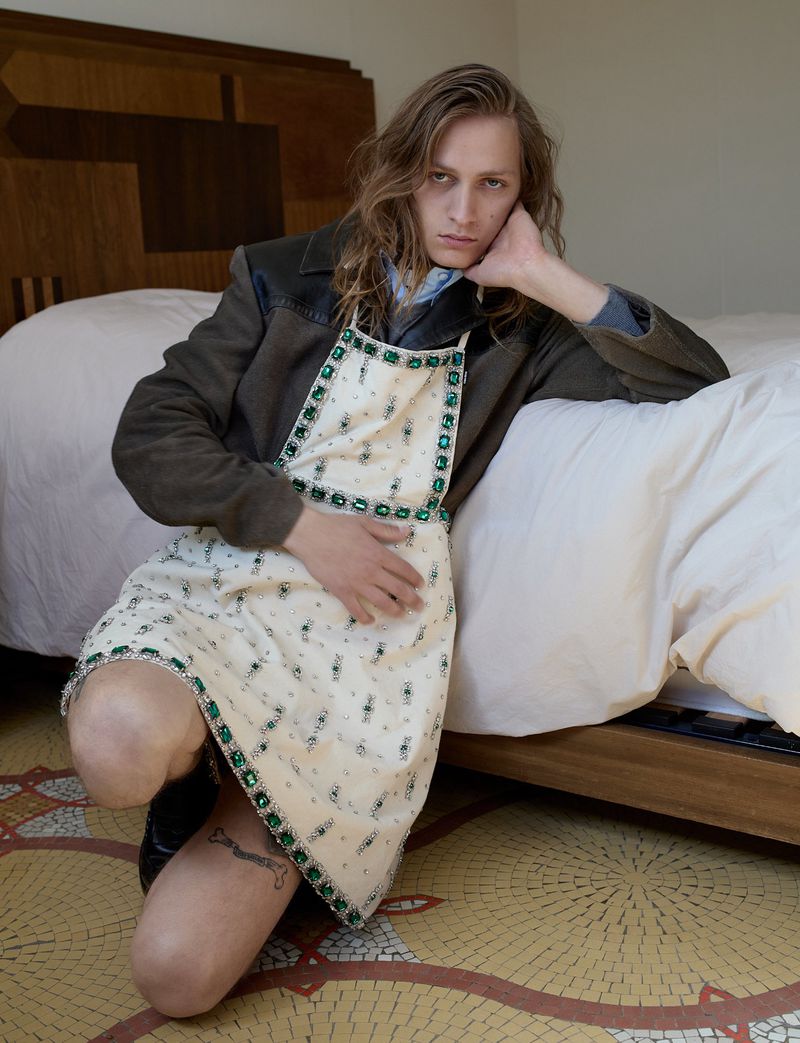 Young man with long hair kneeling beside a bed wearing an embellished cream dress over a dark jacket.