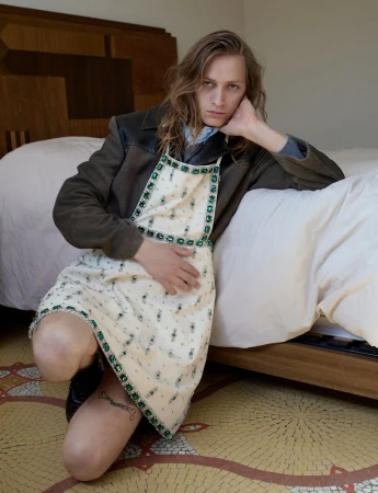 Young man with long hair kneeling beside a bed wearing an embellished cream dress over a dark jacket.