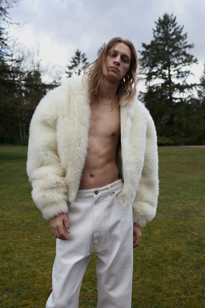 A shirtless male model wearing an open cream fleece jacket and white pants standing outdoors on a grassy lawn with trees.