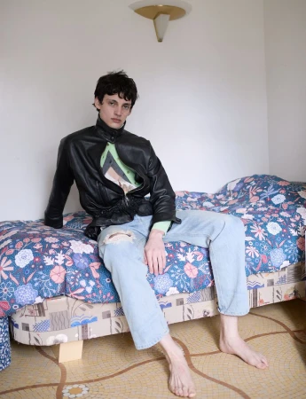 A barefoot male model in a black leather jacket and ripped jeans sits on a floral patterned bed against a white bedroom wall.