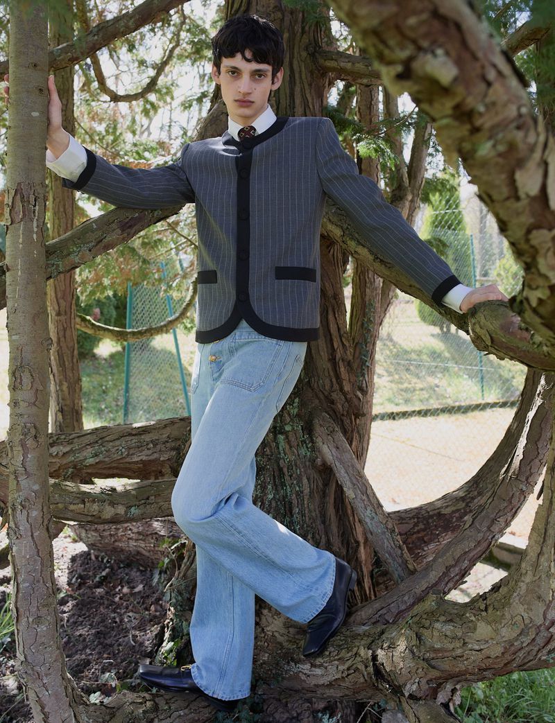 Model posing in the branches of a tree wearing a grey pinstripe jacket with black trim and light wash flared jeans.
