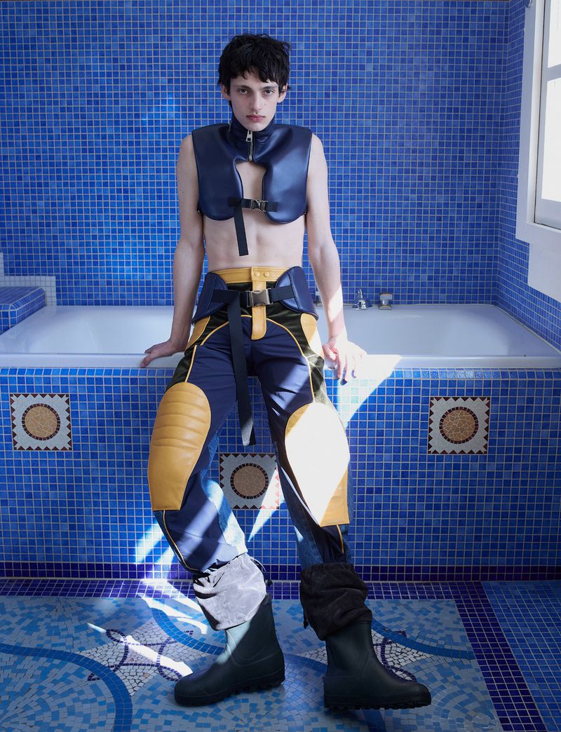 Model wearing dark blue vest and buckled trousers sitting on a tiled bathtub edge in a blue mosaic tiled bathroom.