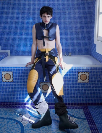 Model wearing dark blue vest and buckled trousers sitting on a tiled bathtub edge in a blue mosaic tiled bathroom.