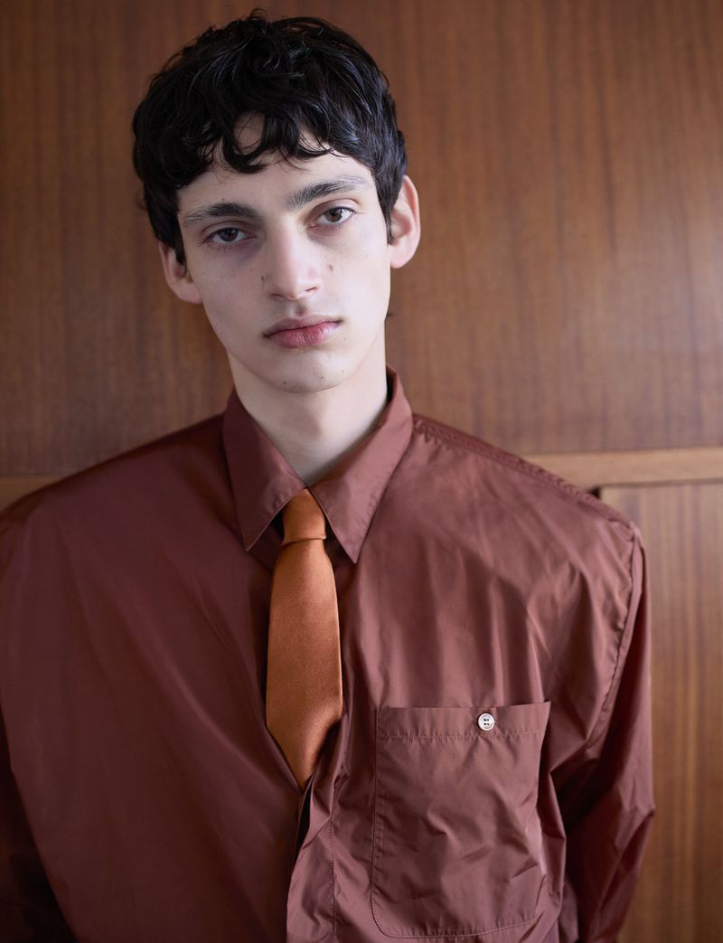 Portrait of a young man wearing a brown button-up shirt and tie against a dark wood paneling background