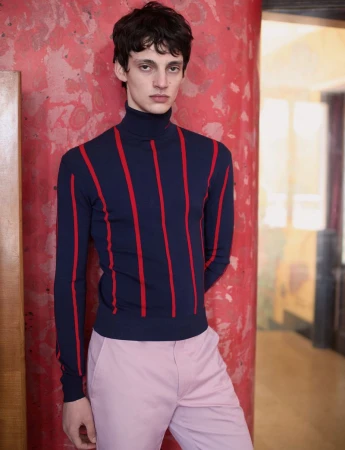Model wearing a navy blue turtleneck with red vertical stripes and pale pink trousers posing against a red textured column.