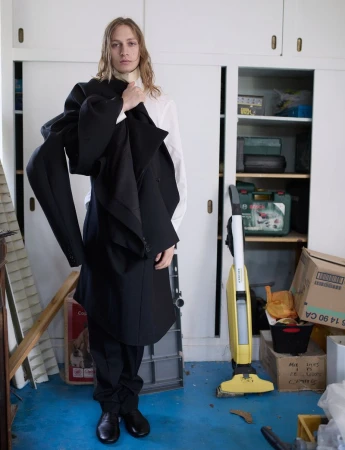 Model wearing a black coat draped over his shoulder in a domestic storage room for Replica Man Magazine