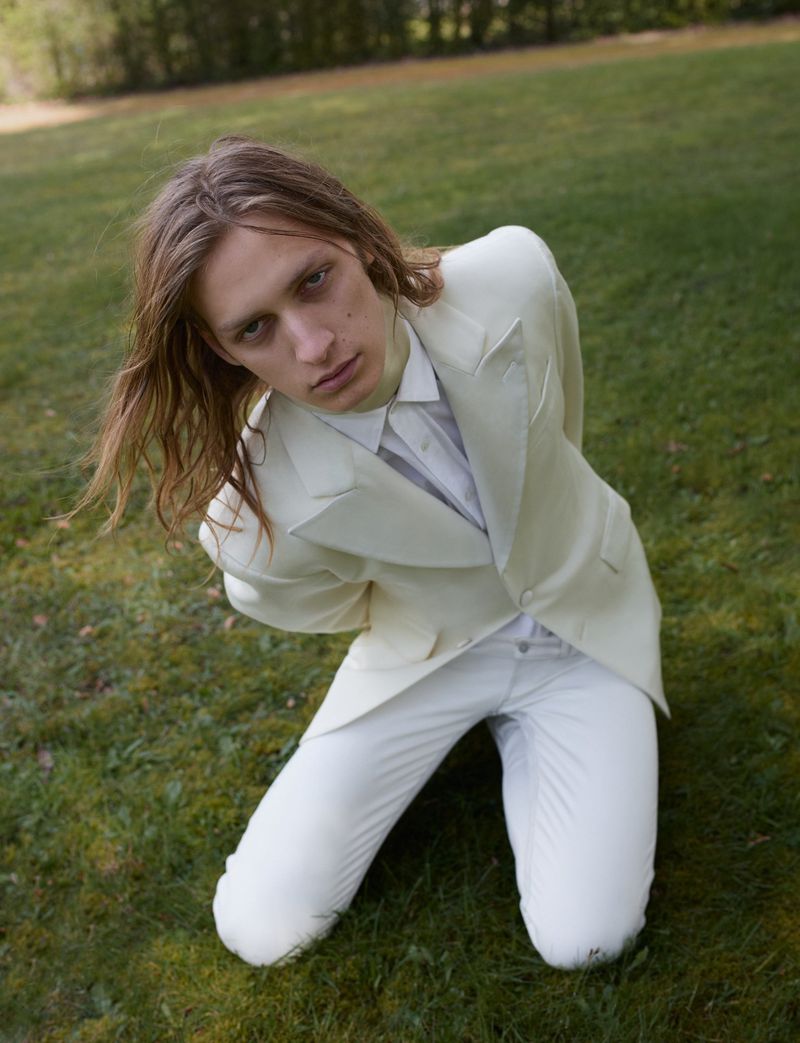 A man with long light brown hair wearing an off-white tailored suit kneeling on a green lawn looking up at the camera.
