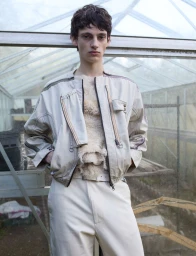 A young male model in a pale beige zippered jacket and trousers posing inside a weathered glass greenhouse.