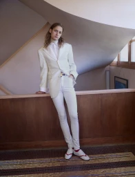 Model in a tailored off-white blazer and white trousers leaning against a wooden balustrade in an architectural interior.
