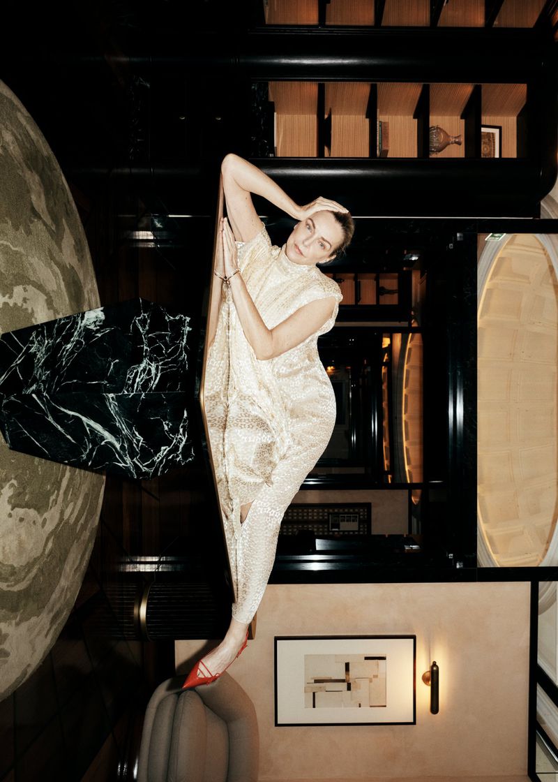 Model Simonetta Gianfelici posing in a shimmering cream sleeveless gown and red pointed-toe heels inside a luxurious interior.