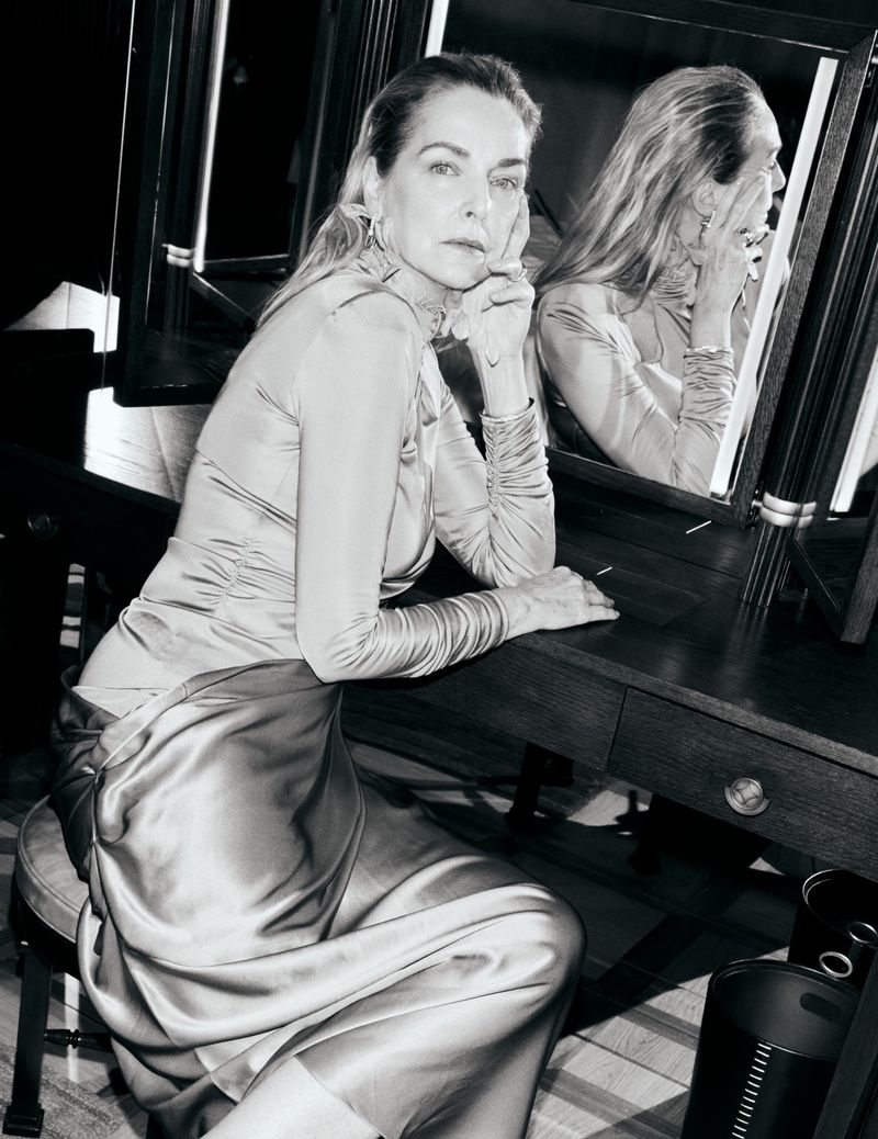 Simonetta Gianfelici wearing a draped satin dress while seated at a dark vanity table with illuminated mirrors in black and white.