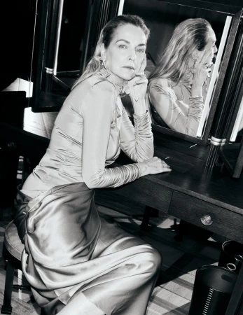 Simonetta Gianfelici wearing a draped satin dress while seated at a dark vanity table with illuminated mirrors in black and white.