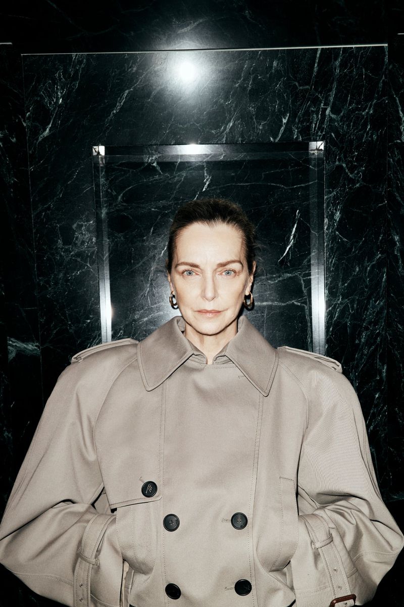 Simonetta Gianfelici wearing an oversized beige double-breasted trench coat in front of a polished dark marble wall.