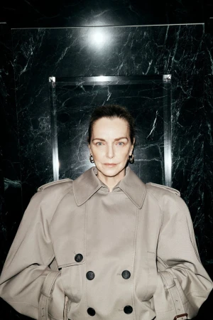 Simonetta Gianfelici wearing an oversized beige double-breasted trench coat in front of a polished dark marble wall.