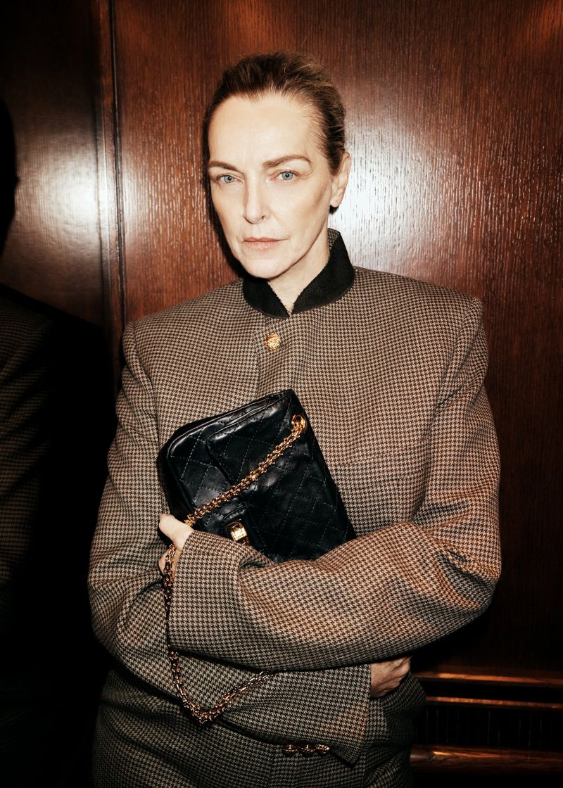 Model Simonetta Gianfelici in a tweed jacket holding a black quilted chain handbag against dark wood paneling.