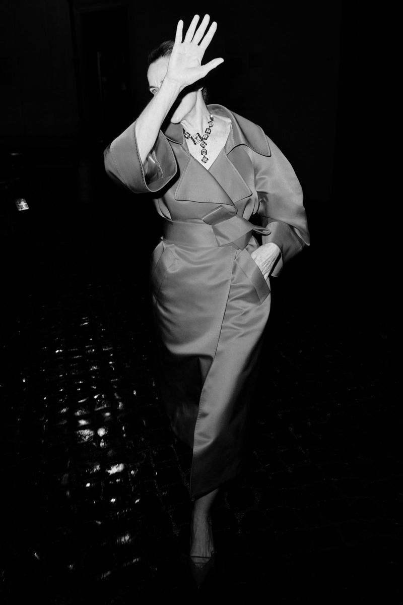 Model Simonetta Gianfelici raises a hand to shield her face while wearing a belted trench coat on a dark cobblestone street.