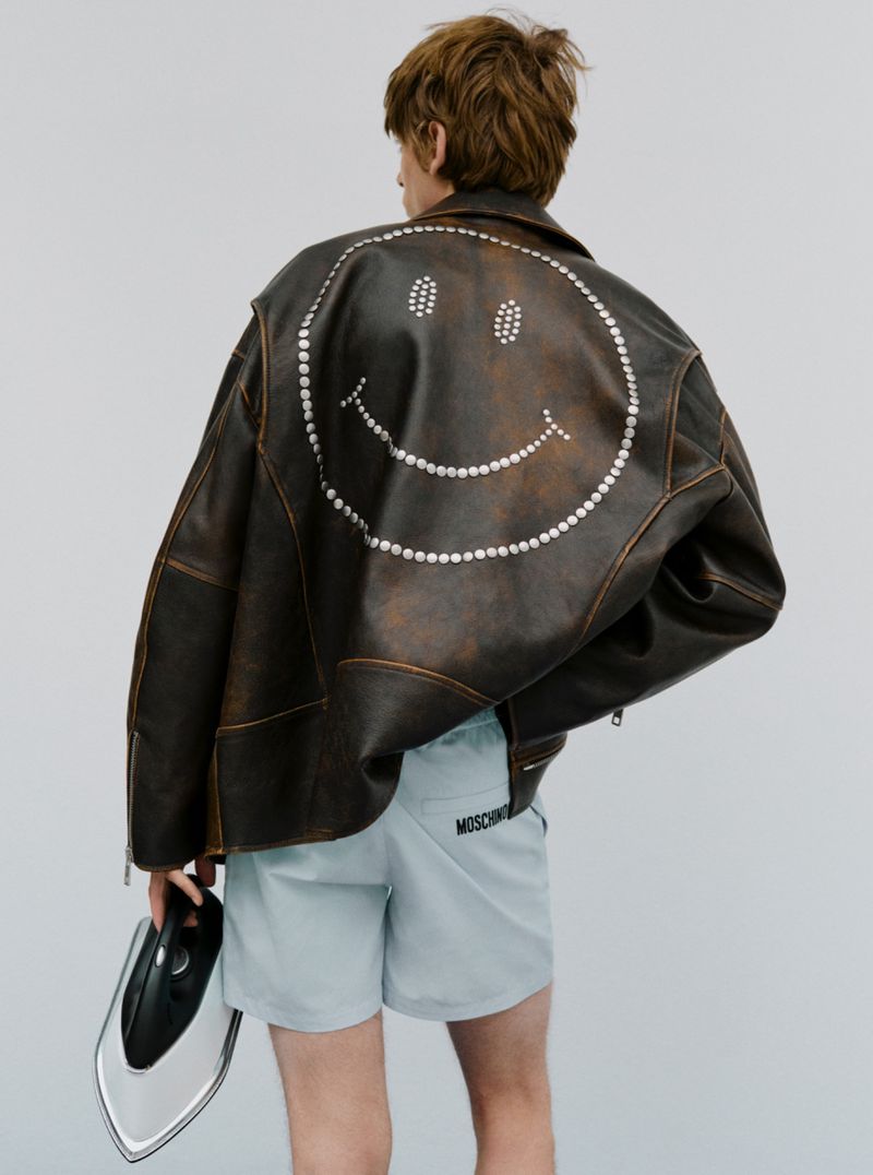 Person from the back wearing a brown leather jacket with a smiley face on the back and blue shorts holding an iron against a white background.