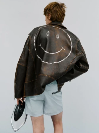 Person from the back wearing a brown leather jacket with a smiley face on the back and blue shorts holding an iron against a white background.
