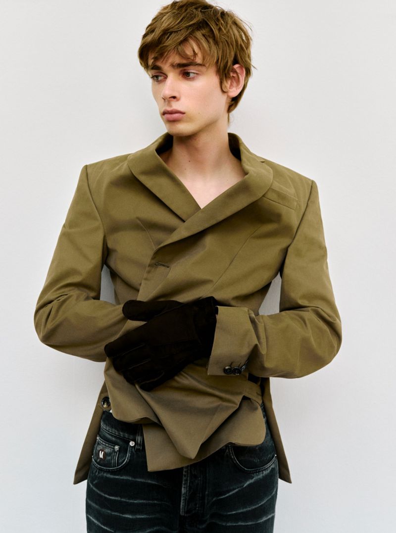 Young adult with light-brown hair in a brown blazer, black gloves, and jeans. The image is a studio shot against a white backdrop.