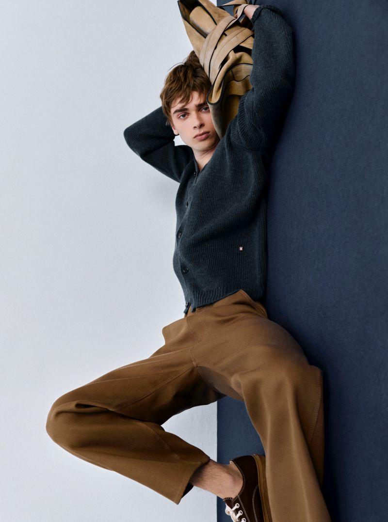Studio shot of a young adult man in a navy cardigan and brown pants leaning against a wall, holding a beige jacket over his shoulder.