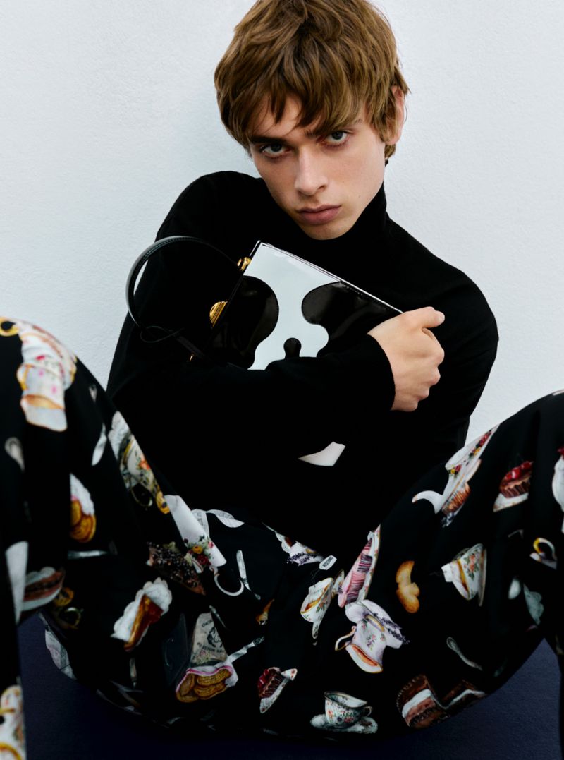 Young adult male with brown hair poses indoors, wearing a black turtleneck sweater and patterned pants, holding a black and white purse.
