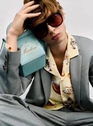 Model in grey suit holding a light blue handbag, wearing tinted sunglasses and printed yellow shirt against a white backdrop.