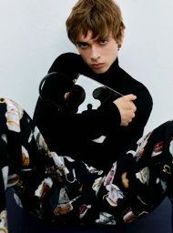 Model wearing a black turtleneck and patterned trousers holding a black and white bag against a white background.
