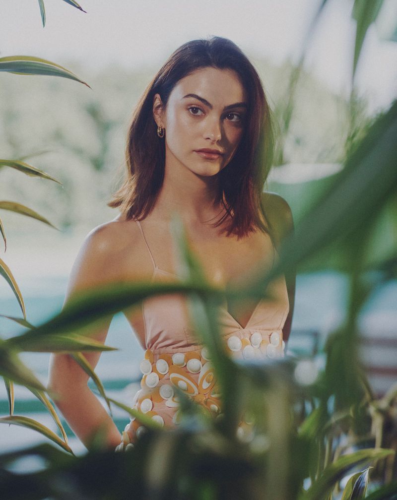Camila Mendes posing in a blush pink dress with floral appliques behind blurred green foliage.