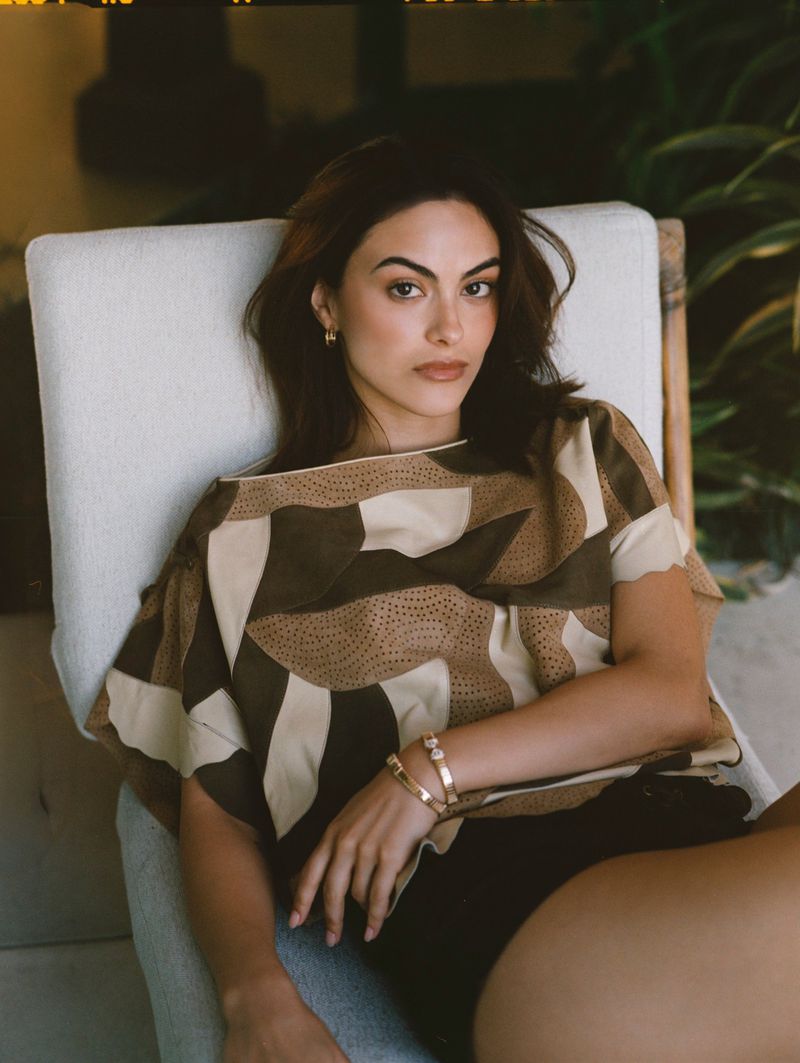 Camila Mendes seated in an off-white cushioned chair wearing a geometric earth-tone top and gold bracelets.