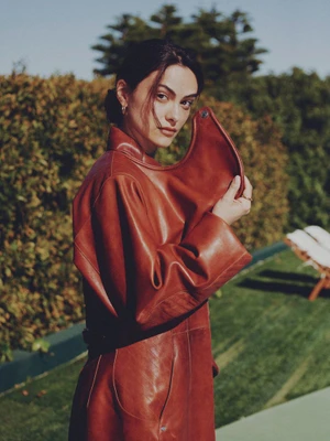 Camila Mendes wearing a structured rust leather coat with an exaggerated collar, posing outdoors beside green hedges.