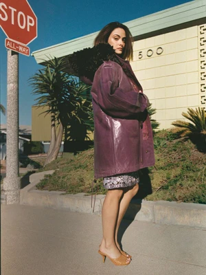 Camila Mendes wearing an oversized purple leather coat with black fur trim on a sunny street sidewalk beside a stop sign.