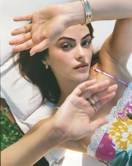 Camila Mendes reclining on an outdoor lounger with raised hands blocking the sun, wearing a floral lace camisole.