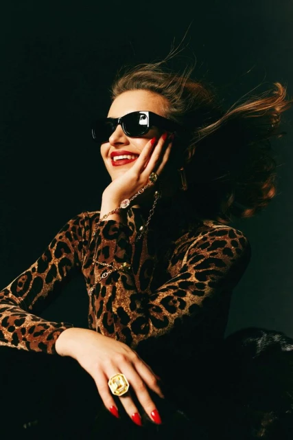 A woman with red lipstick and sunglasses, wearing a leopard print top and gold jewelry, smiles with her hand to her face.