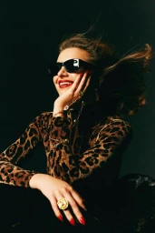 Woman in leopard print top wearing sunglasses and statement rings with red lipstick against dark background