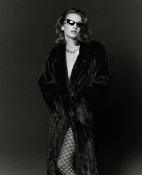 Model in dark sunglasses wearing a black faux fur coat and patterned tights against a dark background