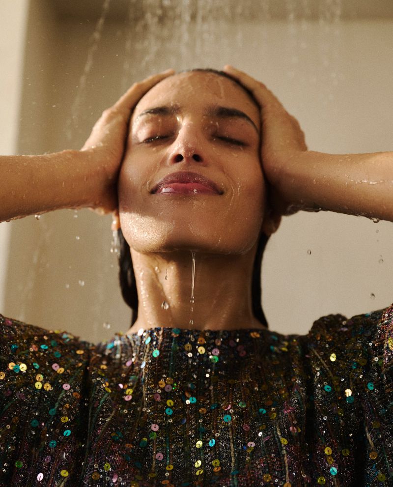 Model Athina Koini with eyes closed and hands to her hair under streaming water, wearing a dark sequined top.