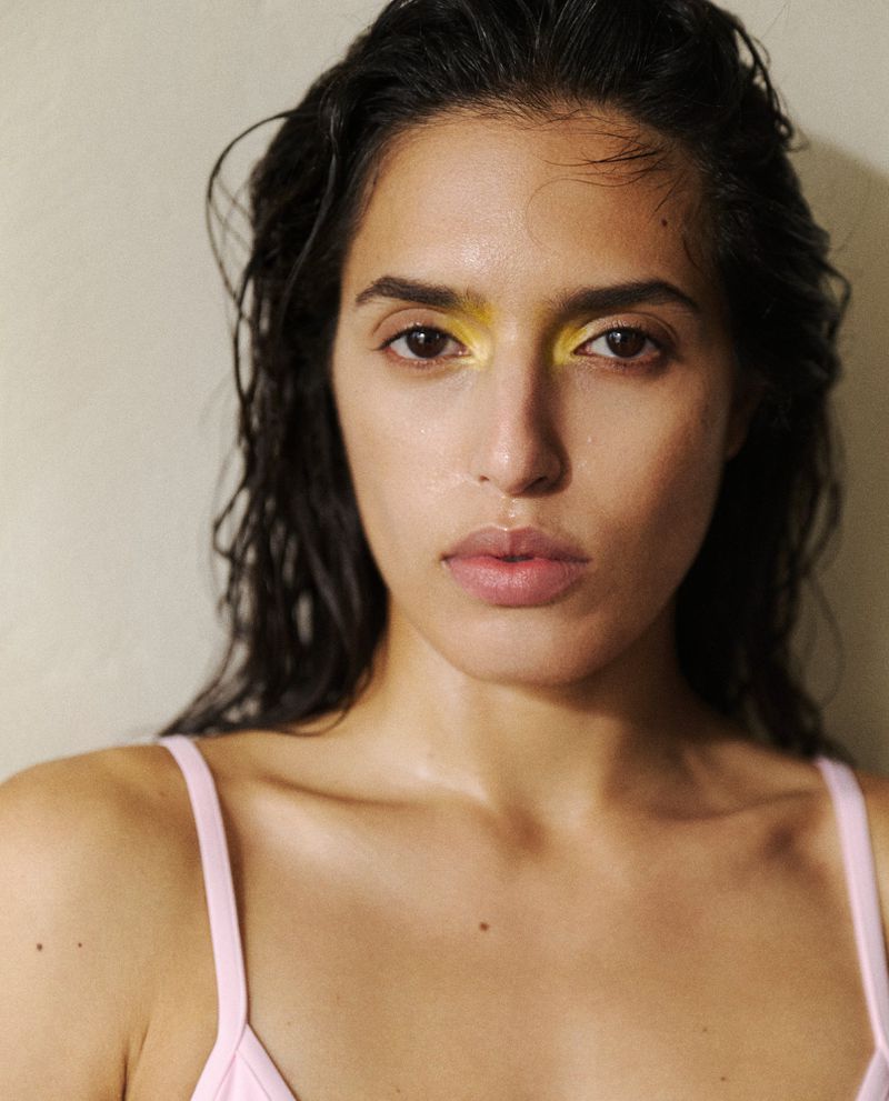 Close-up portrait of model Athina Koini with wet-look hair and vibrant yellow eyeshadow wearing a light pink strap top.