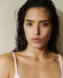 Close-up portrait of model Athina Koini with wet-look hair and vibrant yellow eyeshadow wearing a light pink strap top.