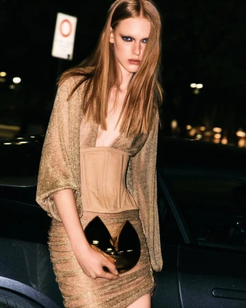 Fashionable woman with red hair in a gold dress and corset stands next to a car on a city street at night, holding a gold clutch purse.