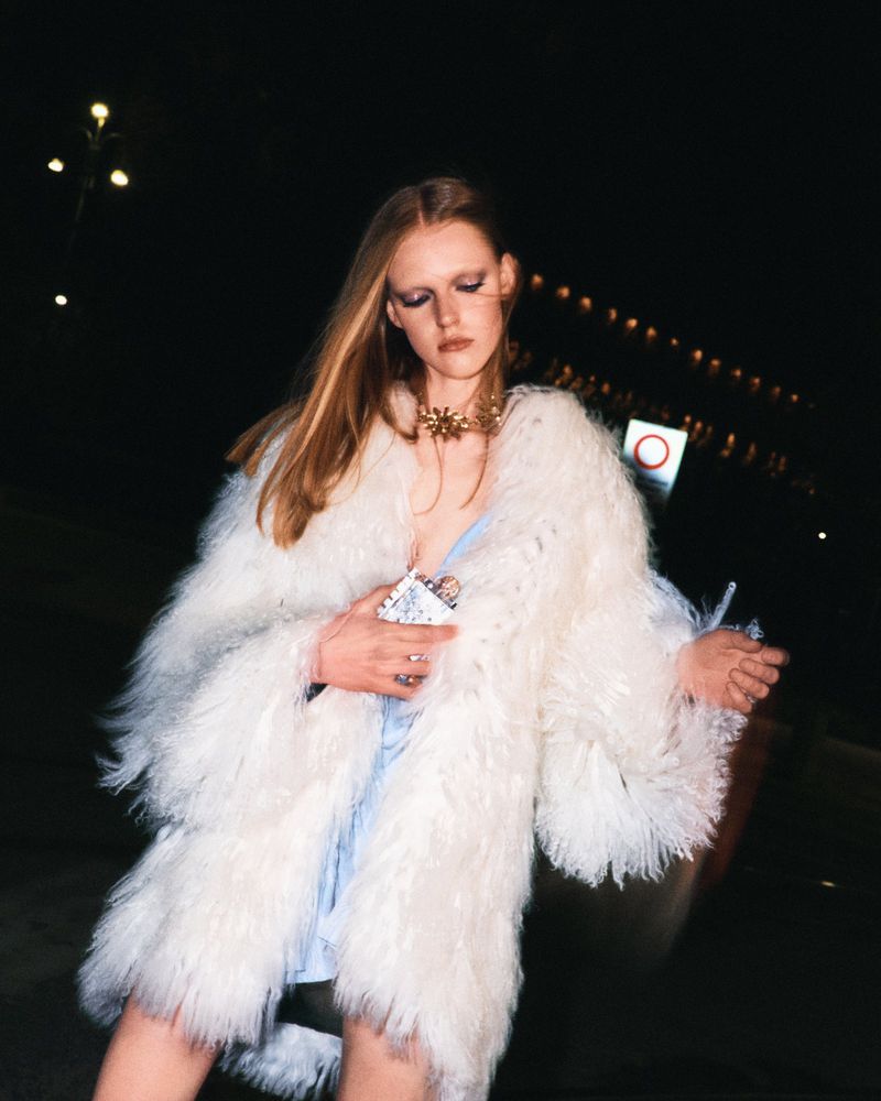 Fashionable woman in a fluffy white coat poses at night. She has long blonde hair and is wearing a gold necklace and a light blue dress.