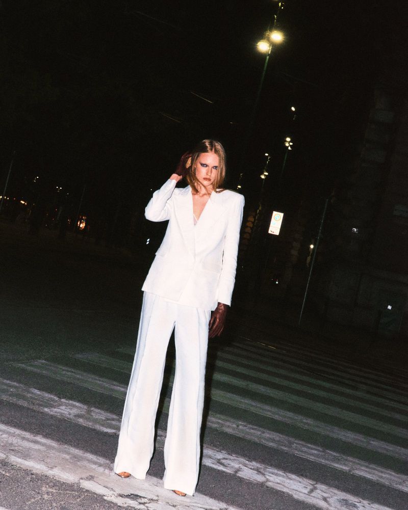 Fashionable young woman in a white suit poses on a city street at night. The crosswalk adds a graphic element to the scene.
