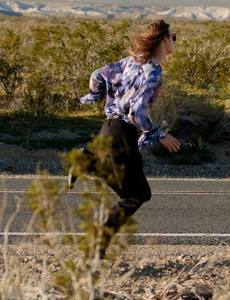 Model Jesse Rinderknecht running across an open desert highway wearing a patterned blue and purple shirt and black trousers.