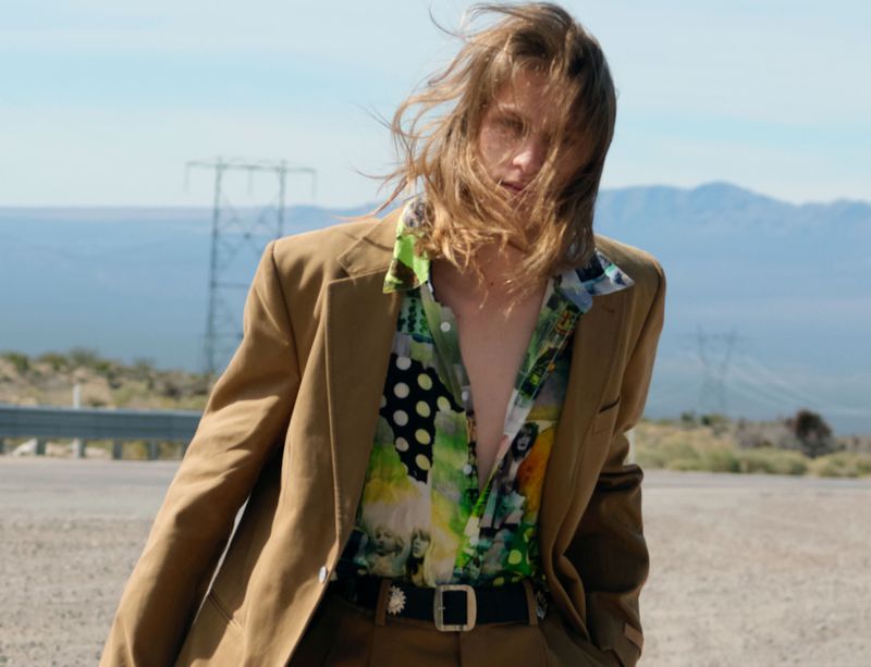 Model with long brown hair wearing a camel blazer and patterned shirt outdoors against a mountainous desert landscape background
