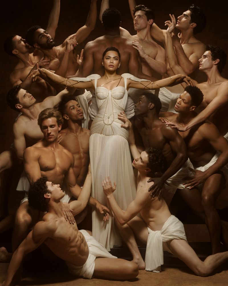 Mona Patel in a sculpted white gown surrounded by shirtless male models draped in white fabric against a warm studio backdrop.