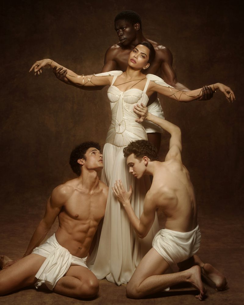 Woman in white gown surrounded by three shirtless male models in classical drapes against a dark textured studio backdrop.