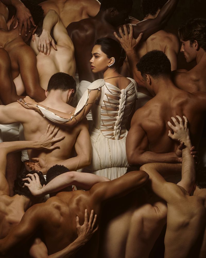 Mona Patel in a sculpted cream gown standing amid shirtless male figures in an editorial fashion portrait.